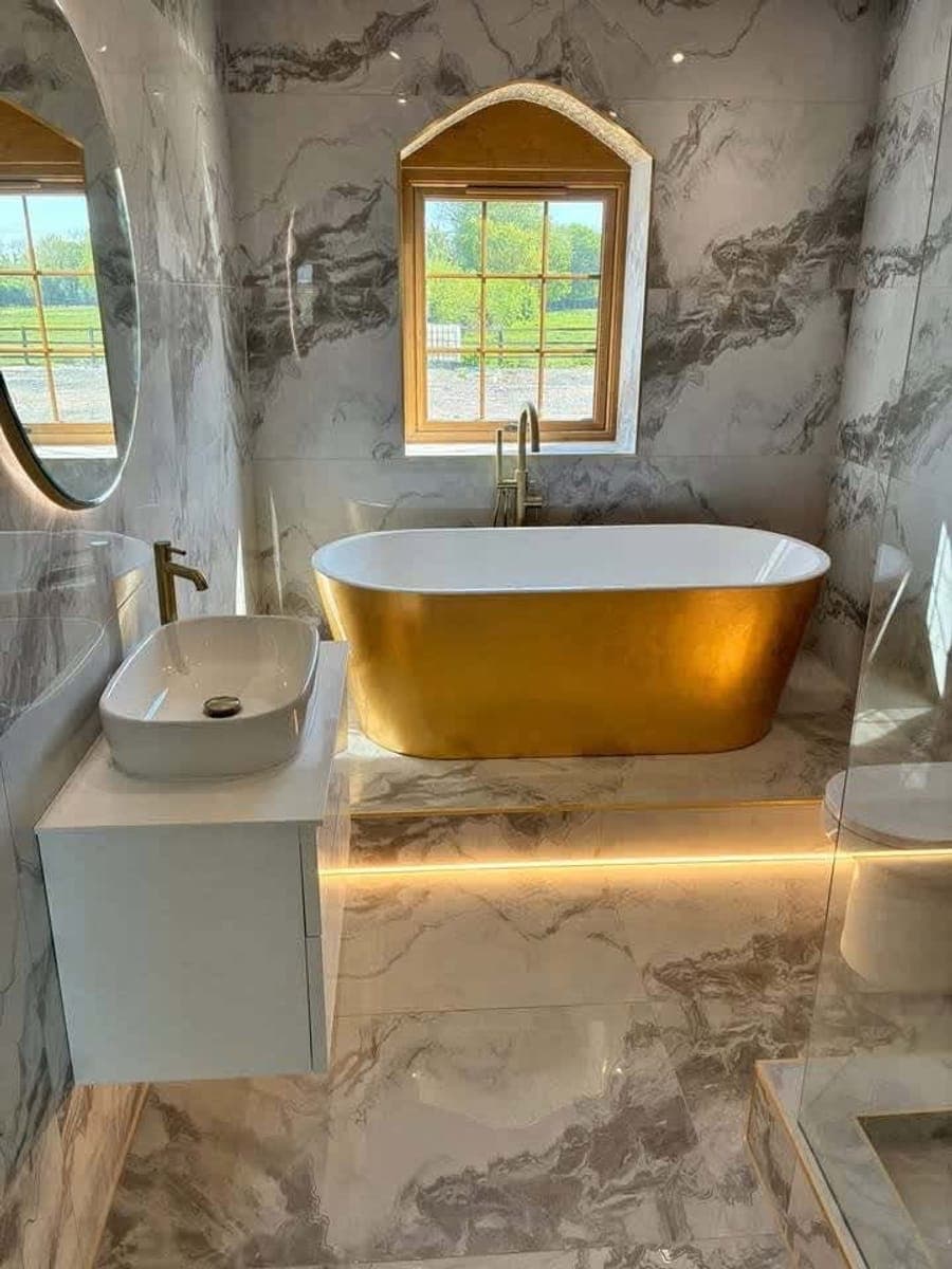 A marble tiled bathroom with a gold leaf freestanding bath and a backlit round mirror