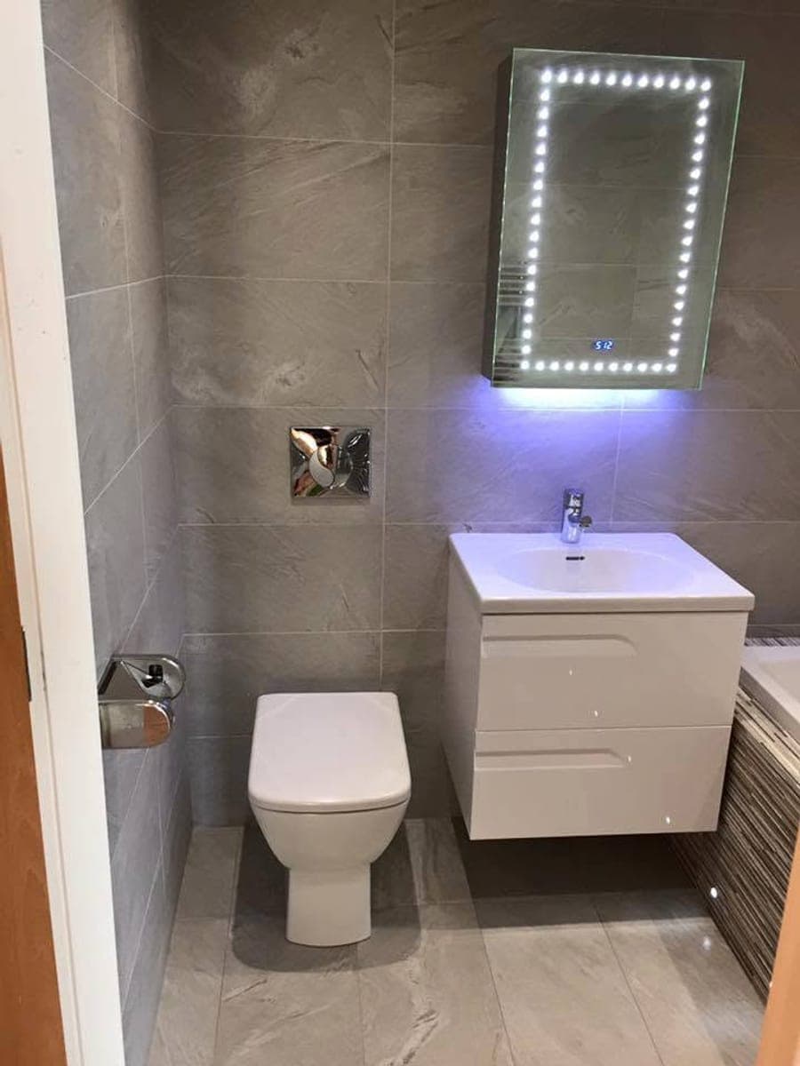 A wall hung toilet beside an LED mirror in a grey tiled bathroom